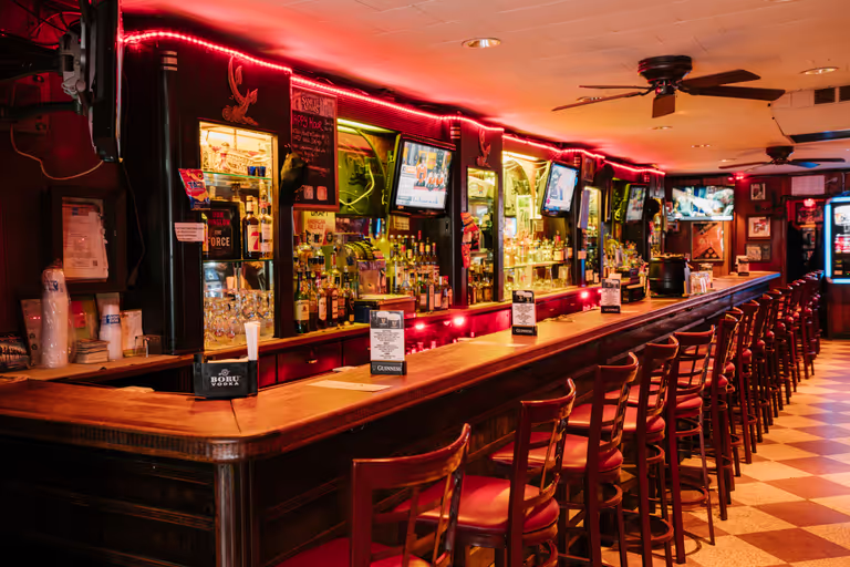 Lengthy bar with several seats, situated under dim neon lighting and ceiling fans. Rows of alcohol line the back wall. 
