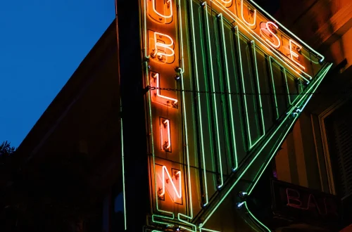 Sign for Dublin House bar. It is a neon green harp, with Dublin spelled vertically along the straight edge. It is neon red. House spelled left to right across the top in neon red. Underneath the harp, the sign includes "Tap Room".