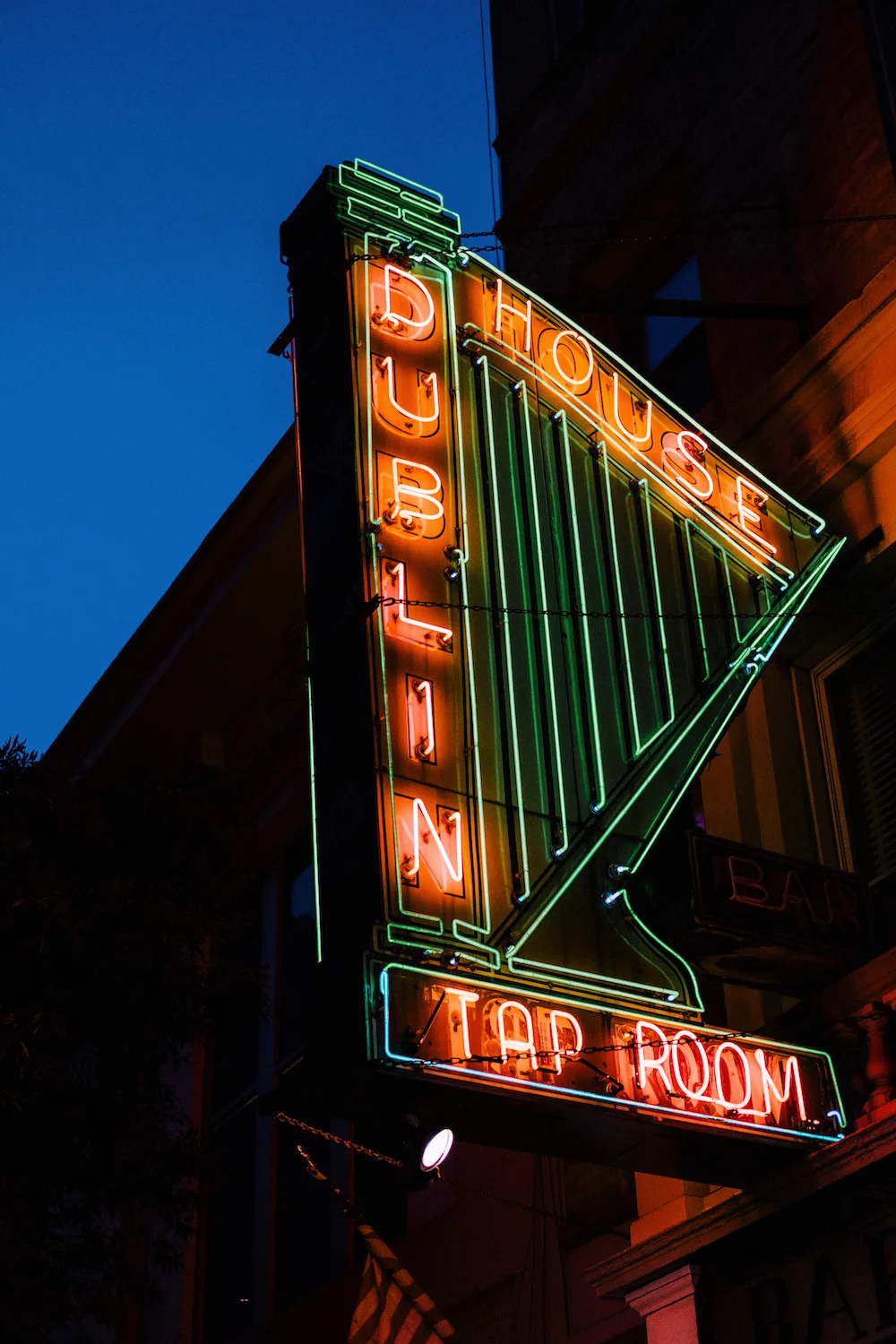 Sign for Dublin House bar. It is a neon green harp, with Dublin spelled vertically along the straight edge. It is neon red. House spelled left to right across the top in neon red. Underneath the harp, the sign includes "Tap Room".